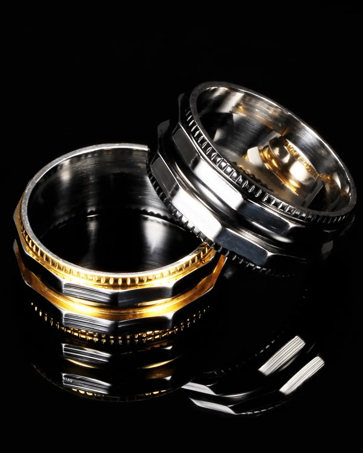 Two metallic rings with a silver and gold color combination, displayed on a reflective black surface against a black background.