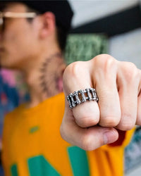A close-up of a person's clenched fist wearing a chunky silver-toned metal ring with a segmented, chain-link design. The background is heavily blurred, showing a person in a yellow and green shirt.
