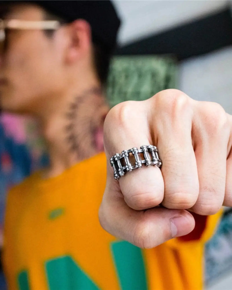 A close-up of a person's clenched fist wearing a chunky silver-toned metal ring with a segmented, chain-link design. The background is heavily blurred, showing a person in a yellow and green shirt.