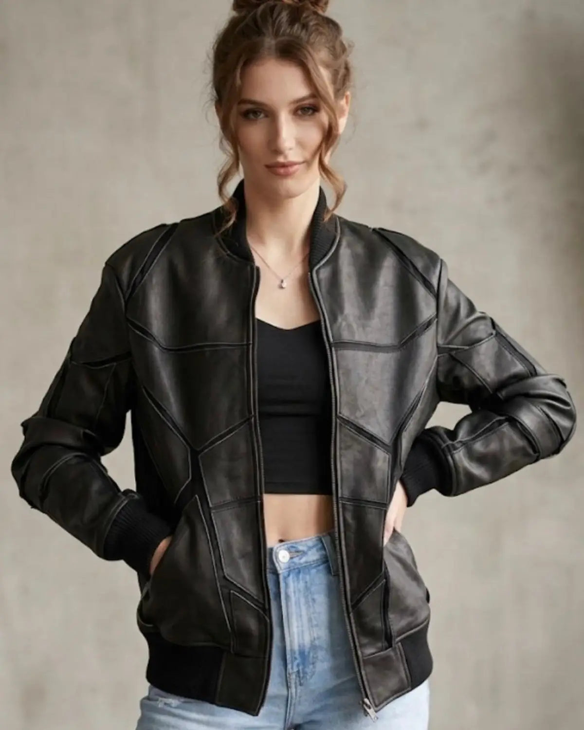 A woman wearing a black leather jacket with geometric paneling, styled over a black crop top and light blue jeans, posing against a textured gray wall.