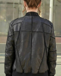 A person viewed from the back wearing a black leather jacket with geometric paneling and contrast stitching, set against a blurred outdoor background.