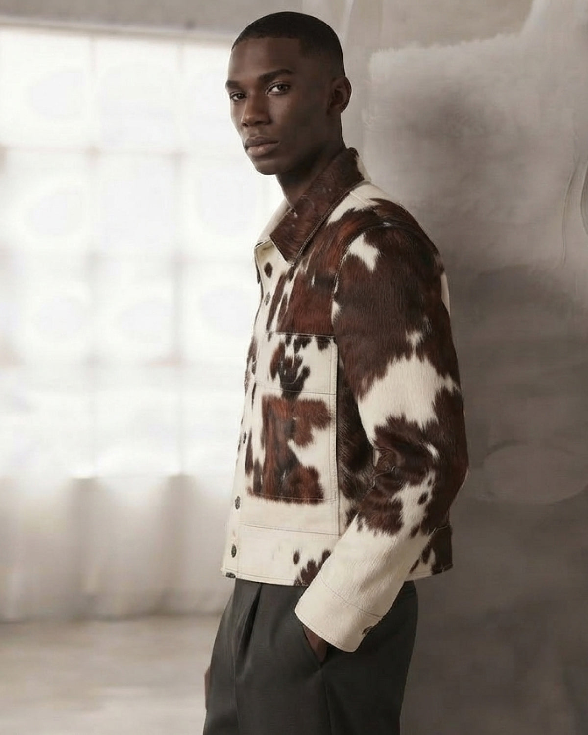 A man wearing a brown and cream cow-print jacket and dark trousers, standing in a softly lit studio setting.