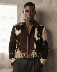 A man wearing a brown and cream cow-print zip-up jacket with dark gray trousers, standing against a textured light gray wall.