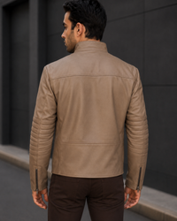 Man wearing a beige leather jacket standing against a dark wall.