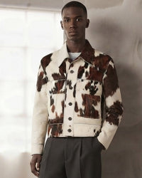 A male model wearing a cropped denim jacket with a brown and cream cow print pattern, paired with dark gray trousers, standing against a textured light gray wall.