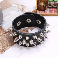 A wide black leather bracelet or cuff decorated with multiple silver metal spikes and oval eyelets, photographed on a tabletop with a blurred decorative background.