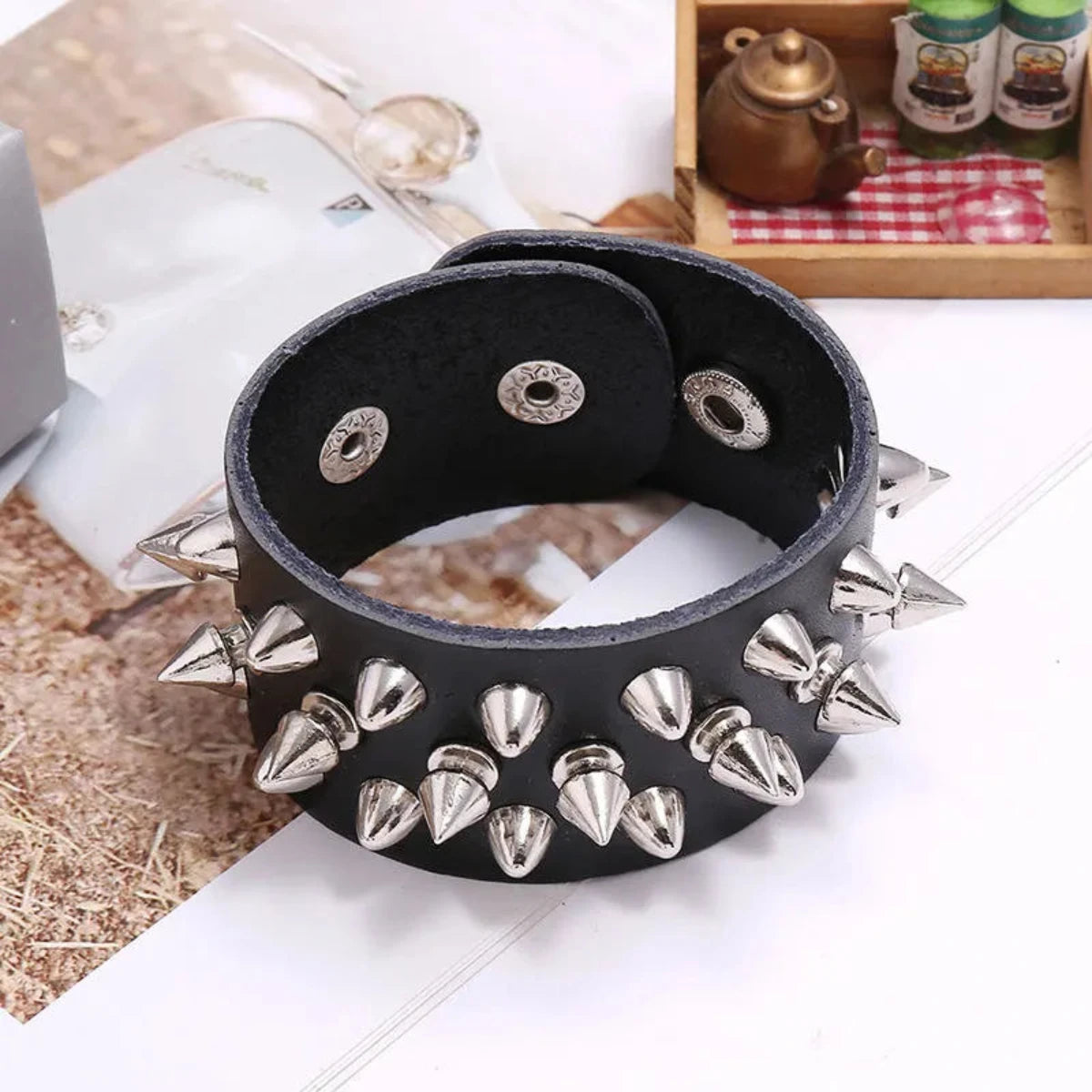 A wide black leather bracelet or cuff decorated with multiple silver metal spikes and oval eyelets, photographed on a tabletop with a blurred decorative background.