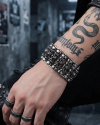 A close-up of a person's tattooed forearm and hand wearing a wide, chunky silver-toned metal bracelet with skull and chain details, along with matching rings, against a dark urban background.