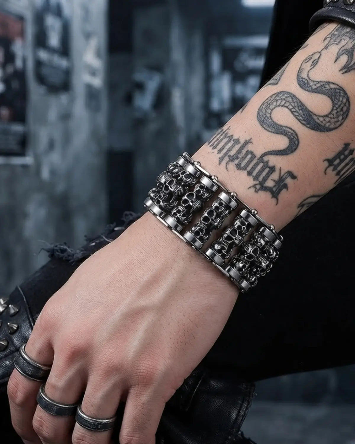 A close-up of a person's tattooed forearm and hand wearing a wide, chunky silver-toned metal bracelet with skull and chain details, along with matching rings, against a dark urban background.