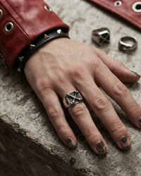 A close-up of a hand wearing a silver geometric ring, resting on a textured stone surface next to a red leather cuff with metal grommets.