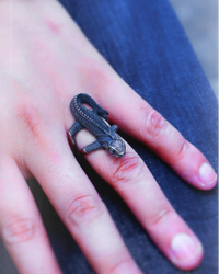 A close-up of a hand wearing a detailed dark metal ring shaped like a small lizard or dragon, resting on dark blue denim fabric.