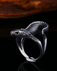 A silver ring sculpted in the shape of a detailed lizard, displayed on a reflective black surface with a blurred brown background.
