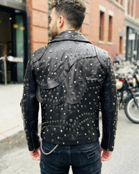A man wearing a black leather jacket heavily embellished with silver studs and eyelets, paired with dark jeans in an outdoor urban setting.