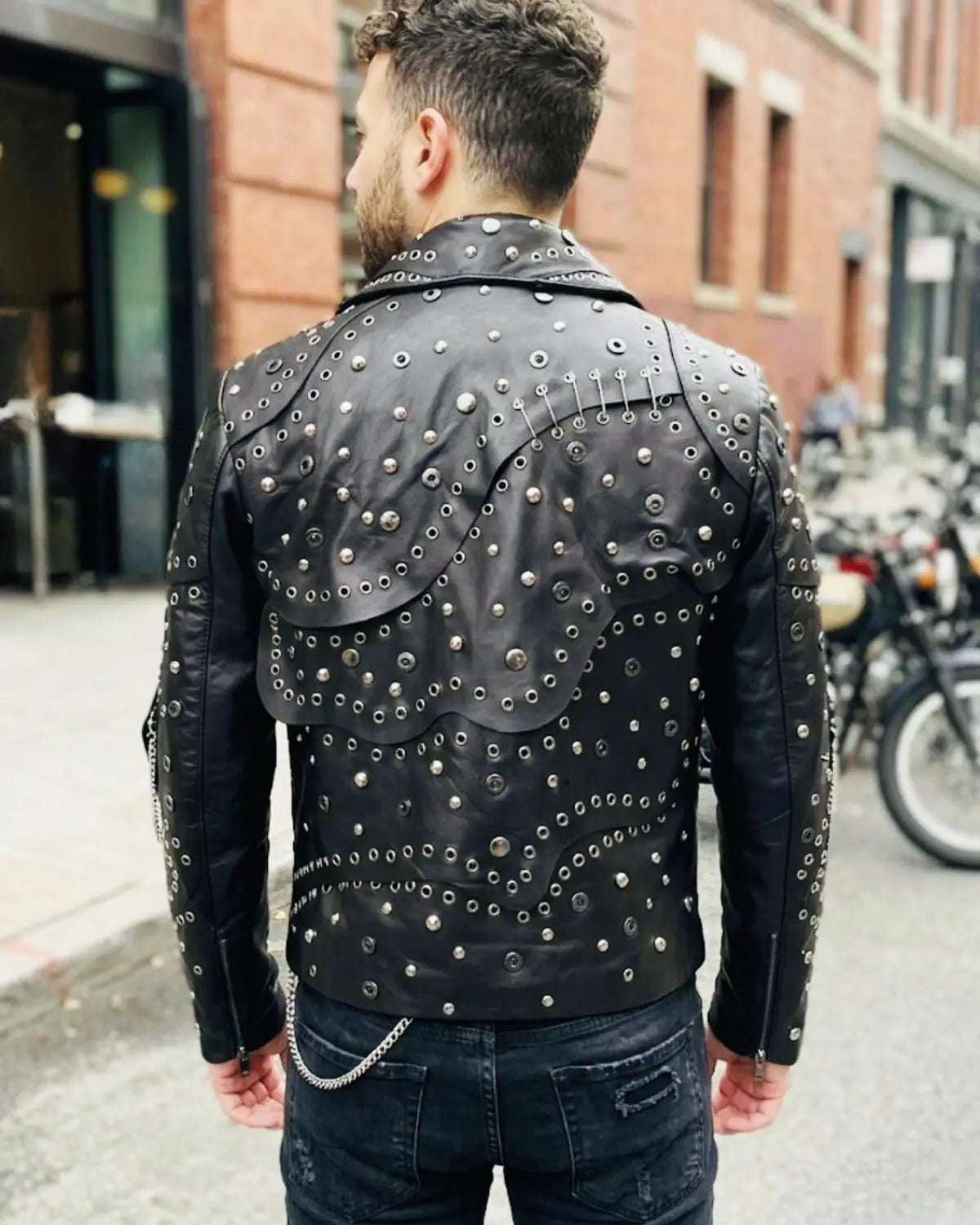 A man wearing a black leather jacket heavily embellished with silver studs and eyelets, paired with dark jeans in an outdoor urban setting.