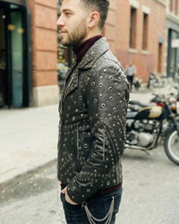 Man wearing a studded leather jacket on a city street with motorcycles in the background.