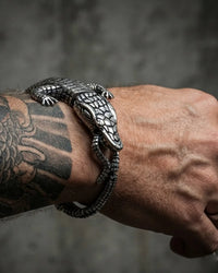 A close-up of a tattooed wrist wearing a detailed silver metal bracelet shaped like a realistic alligator or lizard, set against a dark textured gray background.