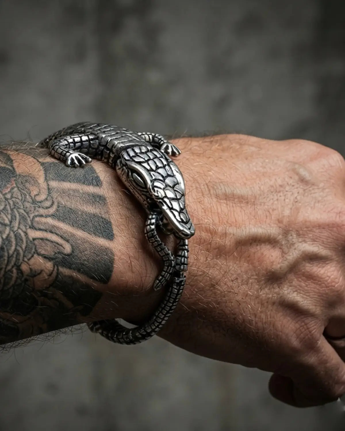A close-up of a tattooed wrist wearing a detailed silver metal bracelet shaped like a realistic alligator or lizard, set against a dark textured gray background.