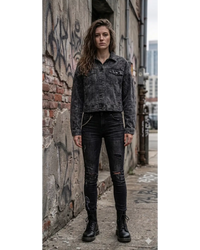 A woman stands on a city sidewalk wearing a faded black washed denim jacket, black distressed skinny jeans, and black combat boots, posed against a weathered brick wall with graffiti.