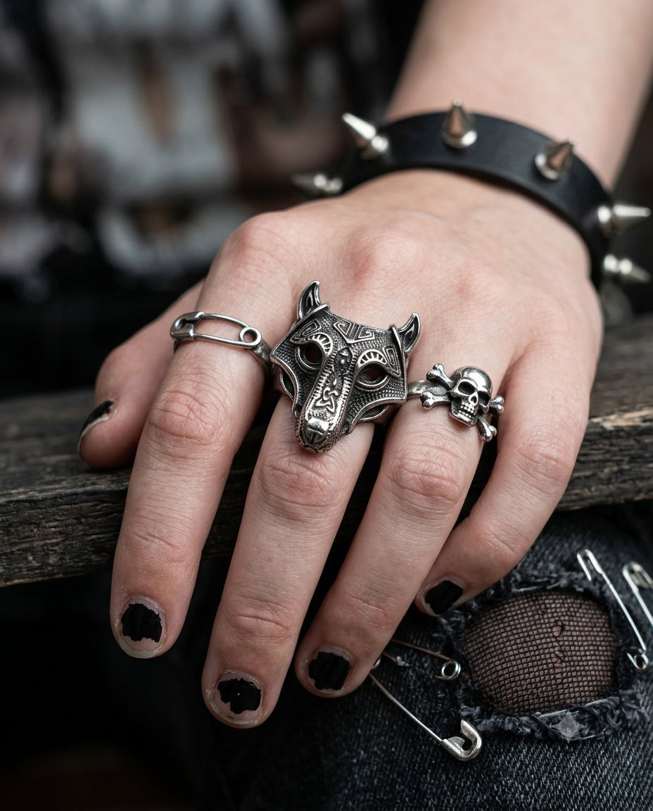 A close-up of a hand wearing multiple silver-toned gothic rings, including a large wolf head ring, a skull ring, and a safety-pin ring, along with a spiked black leather wristband.