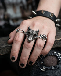 A close-up of a hand wearing multiple silver-toned gothic rings, including a large wolf head ring, a skull ring, and a safety-pin ring, along with a spiked black leather wristband.
