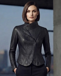 A woman modeling a black leather jacket with a high mock neck, asymmetrical wrap front, and textured woven panel detailing, paired with black trousers against a blurred urban background.
