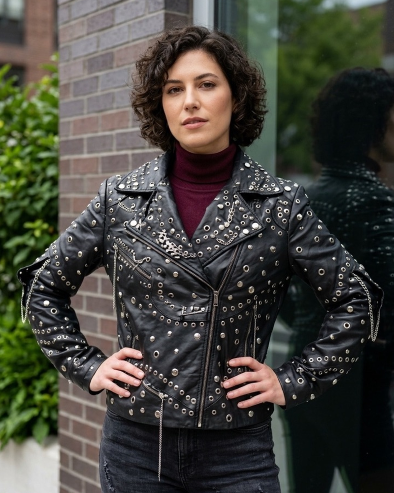 A woman wearing a black leather motorcycle jacket heavily embellished with silver studs and grommets, styled over a dark burgundy turtleneck and black jeans, standing outdoors against a brick wall.