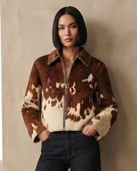 A woman with a short dark bob wears a brown and cream cow-print faux fur jacket with a front zipper, standing against a textured beige wall.