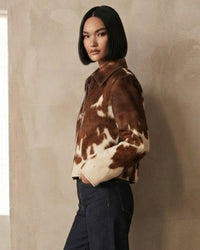 A woman with a short dark bob hairstyle models a brown and cream cow-print suede jacket against a textured beige wall.