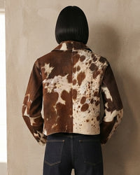 A person shown from the back wearing a cropped cow-print jacket with brown and cream patches, paired with dark blue jeans, standing against a textured beige wall.